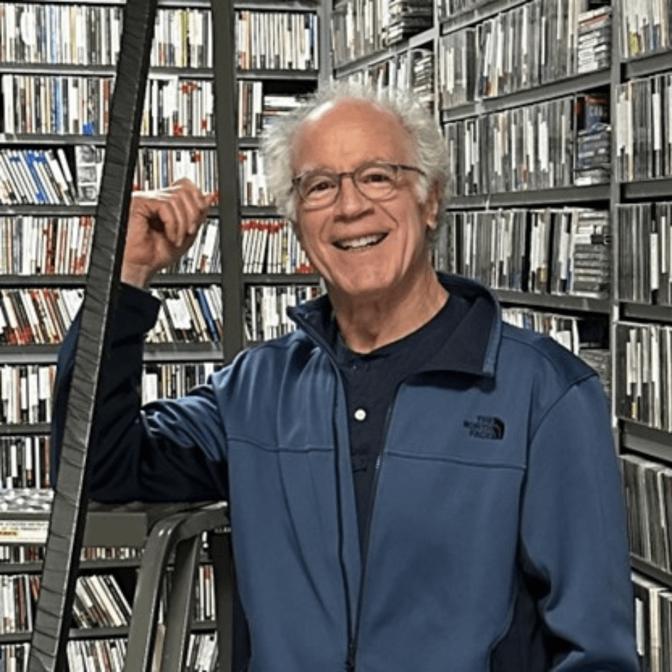 donor Ron Choen stands in a the middle of the photograph in a blue jacket smiling, leaning on a step ladder in a room with wall to wall shelving containing CDs